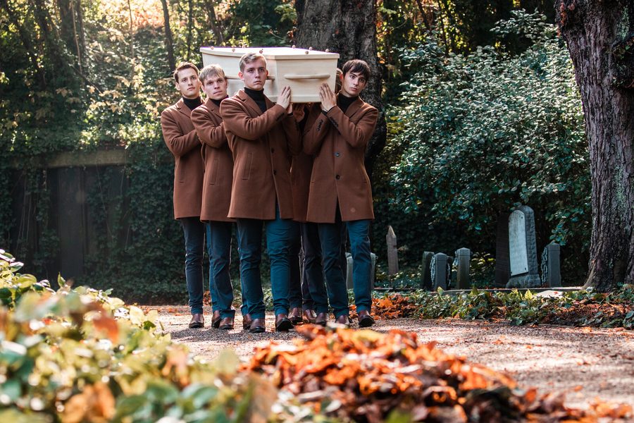 Dragers in bruine jasjes dragen een kist over een begraafplaats