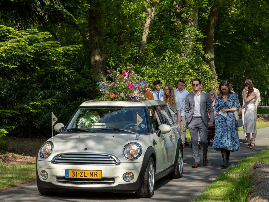 De witte Minirouwauto met een grote bos zomerbloemen op het dak, gevolgd door familie die meeloopt over een bospad