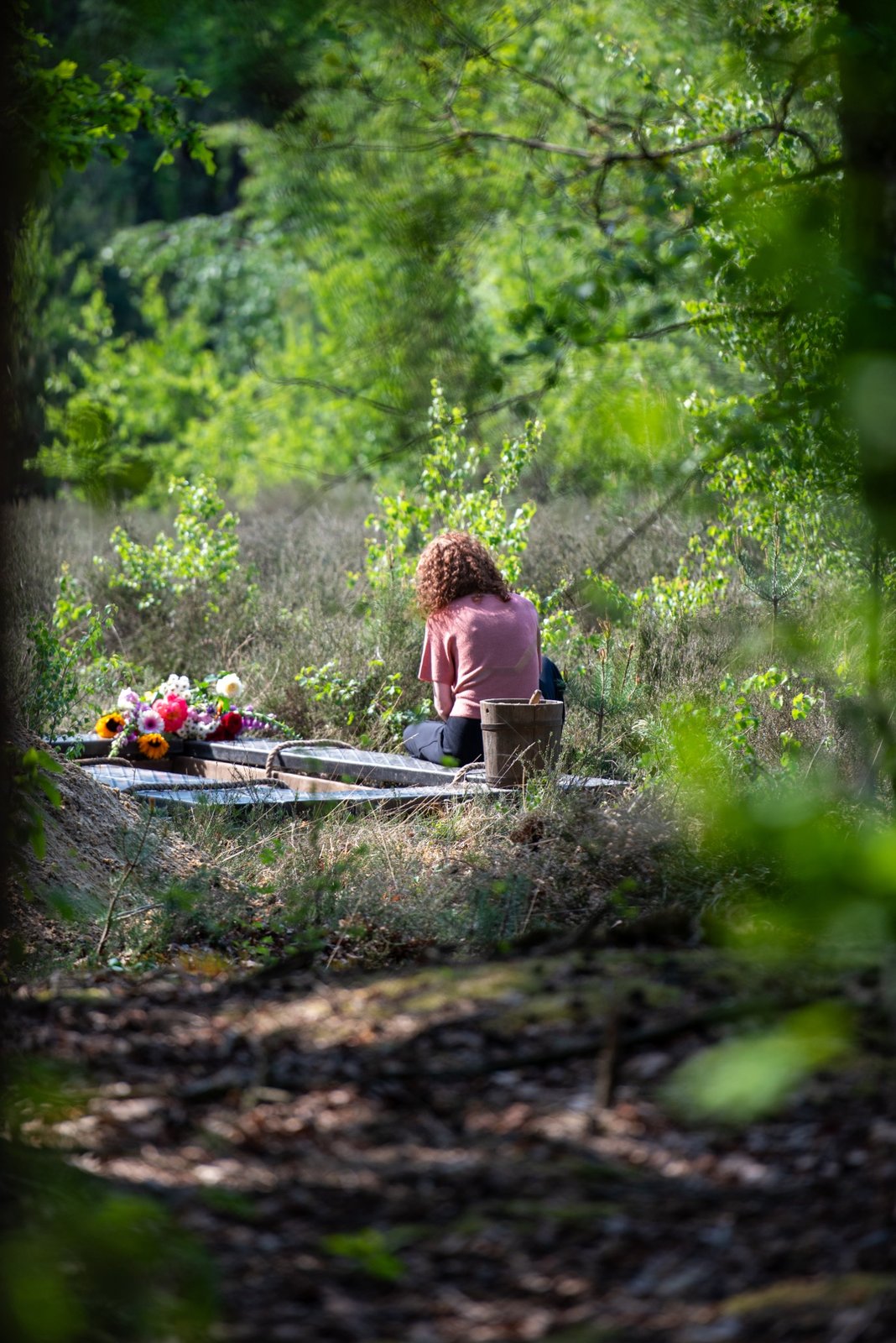 Vrouw zit bij een graf in het bos tussen de heide, met bloemen op het graf