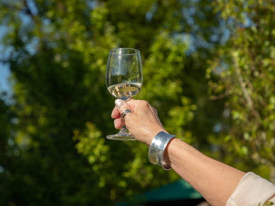 Een hand met een glas witte wijn geheven in een toost, tegen een achtergrond van zonnig groen