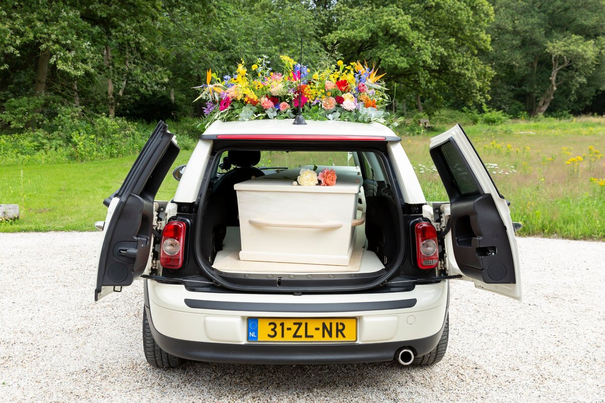 De Minirouwauto met open achterdeuren, de kist erin en bloemen op het dak