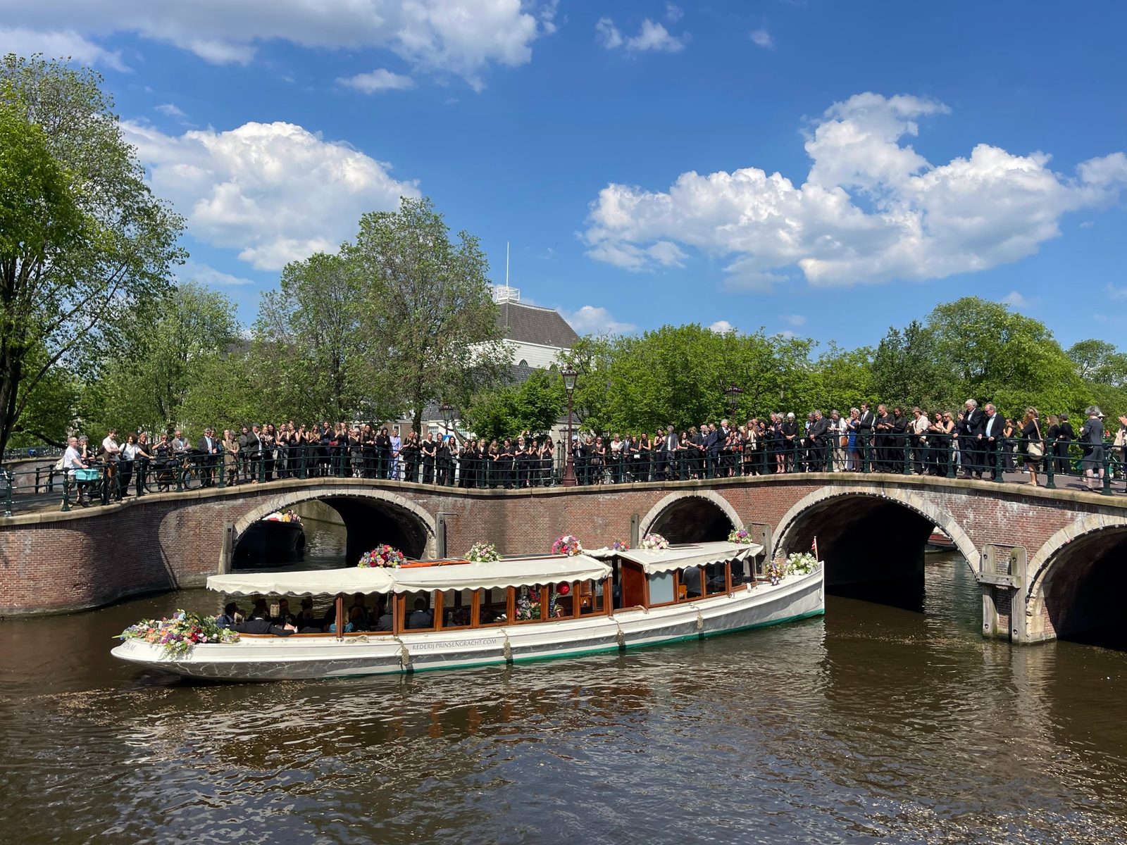 Wateruitvaart in Amsterdam bij De Duif: een salonboot met bloemen vaart over de Prinsengracht onder een brug door, terwijl familie en vrienden op de brug staan